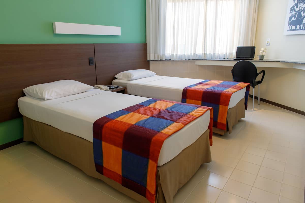 Two single beds with colorful blankets, a desk with a laptop, and a chair in a room with green walls and a window with curtains.