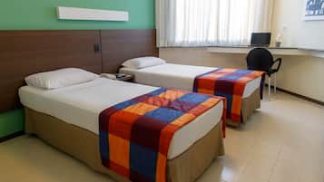 Duas camas de solteiro com cobertores coloridos, uma escrivaninha com um laptop e uma cadeira em um quarto com paredes verdes e uma janela com cortinas.