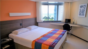 A hotel room with a bed, a desk, and a window with a view of the ocean and buildings. - Park Hotel (Recife)