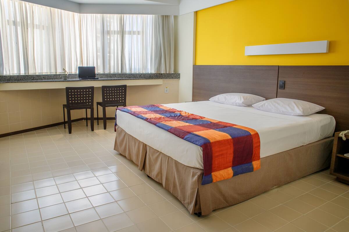 A hotel room with a bed, a desk with a laptop, two chairs, and a window with curtains.