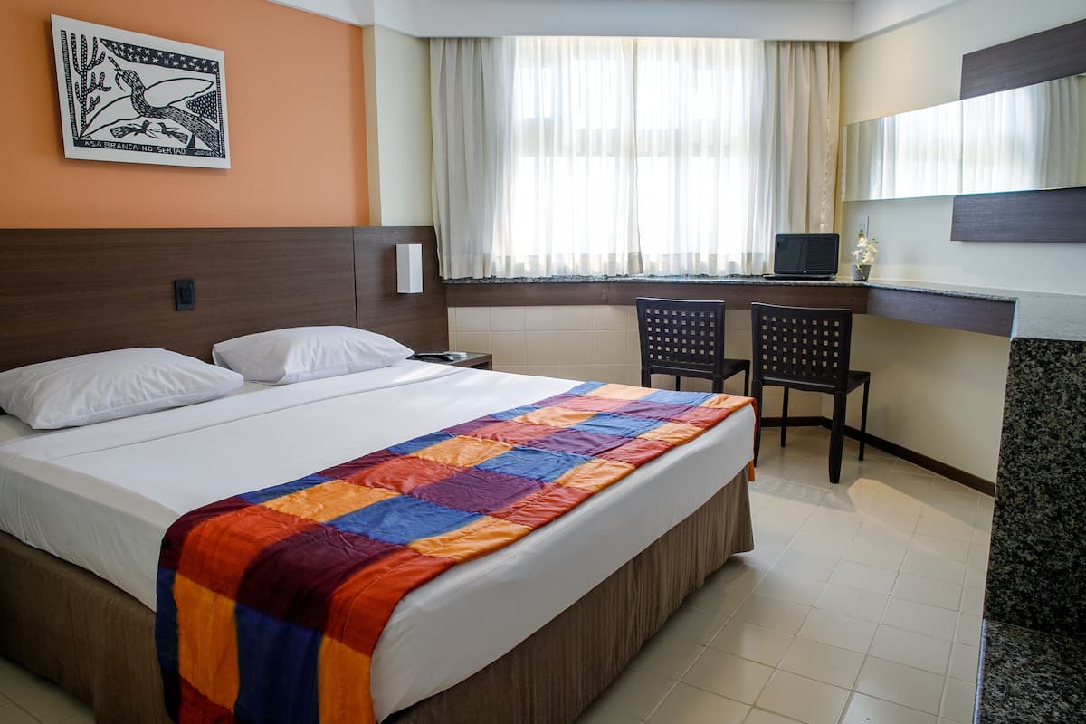 A hotel room with a bed, a desk, two chairs, a window with curtains, a painting on the wall, and a television on the desk.