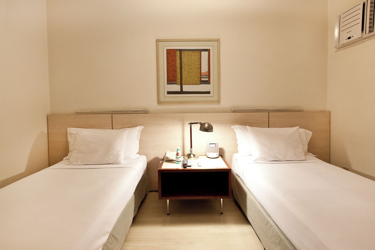 A hotel room with two single beds, a bedside table with a lamp, a phone, and a small table with a bottle and glasses.