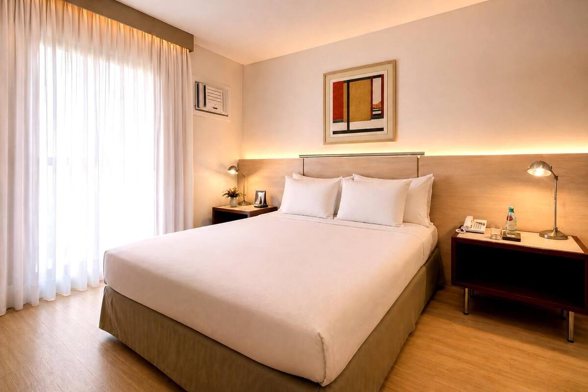 Ein Hotelzimmer mit einem großen Bett, zwei Nachttischen, einem Schreibtisch, einem gerahmten Bild an der Wand und einem Fenster mit Vorhängen.