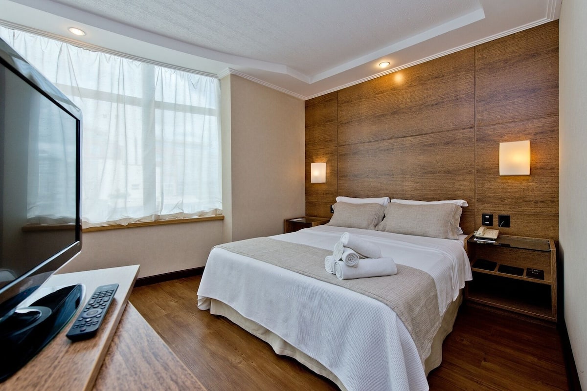 A hotel room with a bed, a TV, a bedside table with a remote control, and a wooden headboard.