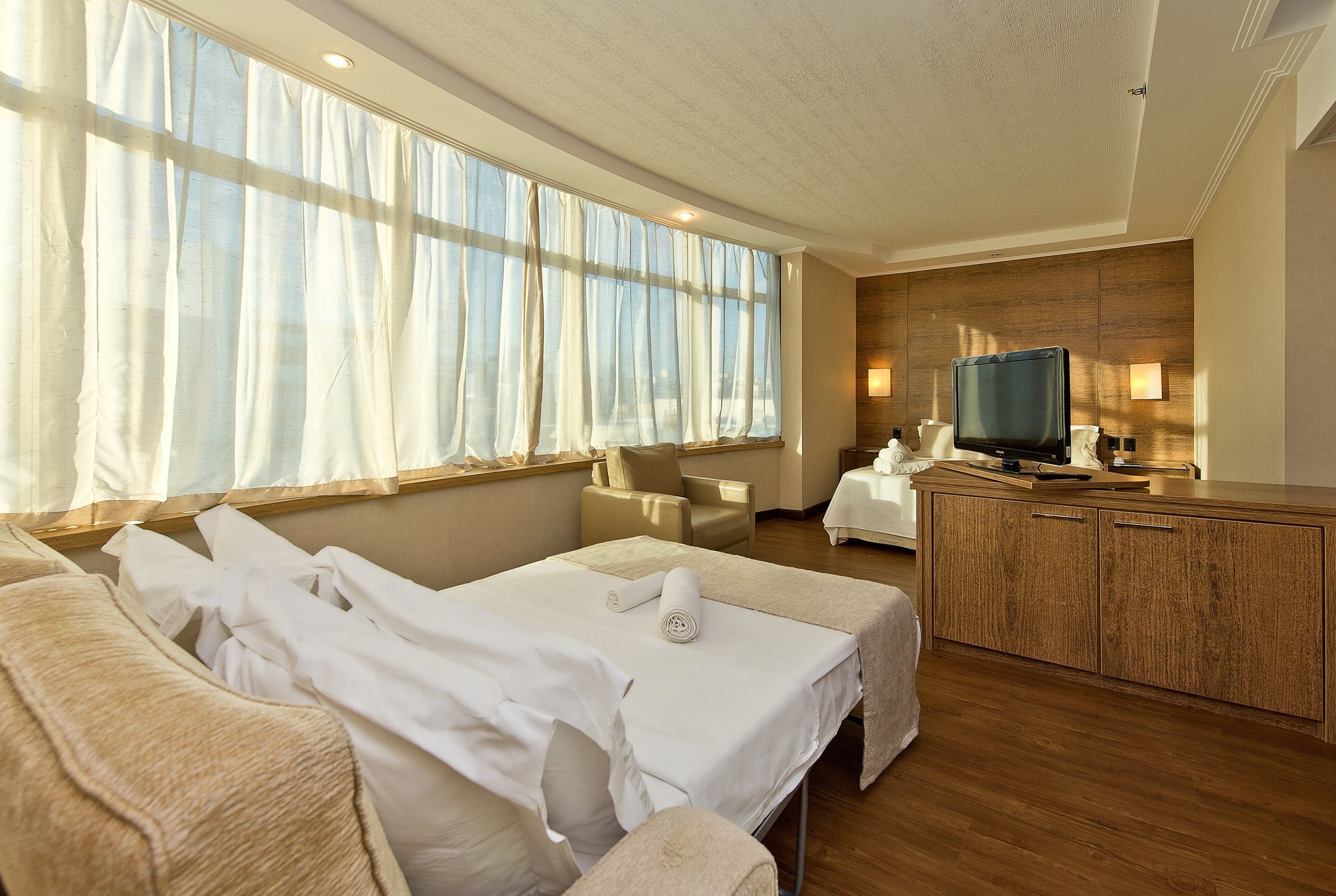 Ein Hotelzimmer mit einem großen Bett, einem Fernseher, einem Holzschrank und einem Fenster mit transparenten Vorhängen.