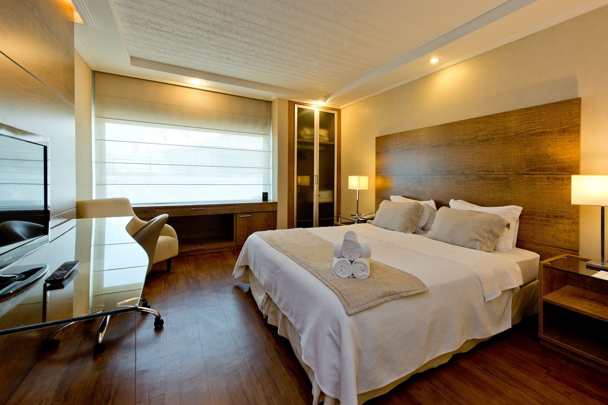 A modern hotel room with a large bed, a desk, and a chair. There is a window with blinds, a wooden headboard, and two bedside lamps.