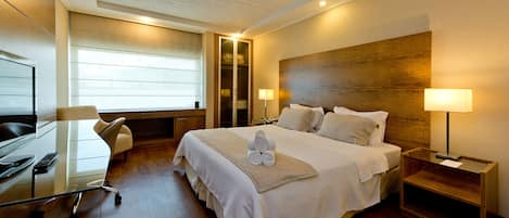 A modern hotel room with a large bed, a desk, and a chair. There is a window with blinds, a wooden headboard, and two bedside lamps.