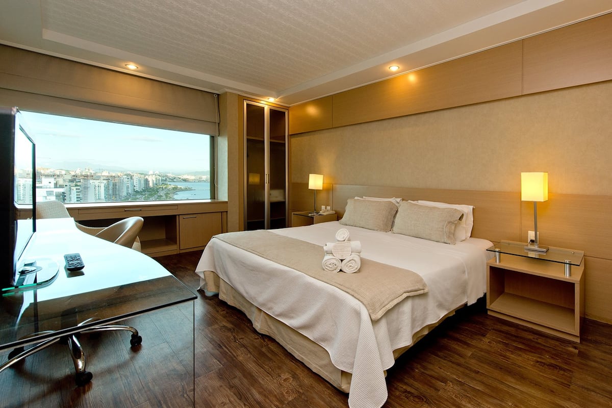 A modern hotel room with a large bed, a desk with a computer, and a view of the city.