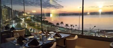 A dining area with a table set for two, overlooking a waterfront with a cityscape in the background.
