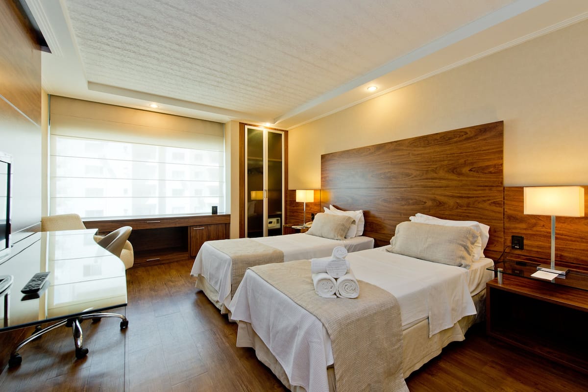 A hotel room with two beds, a desk, a chair, a TV, and a window with a view of buildings.