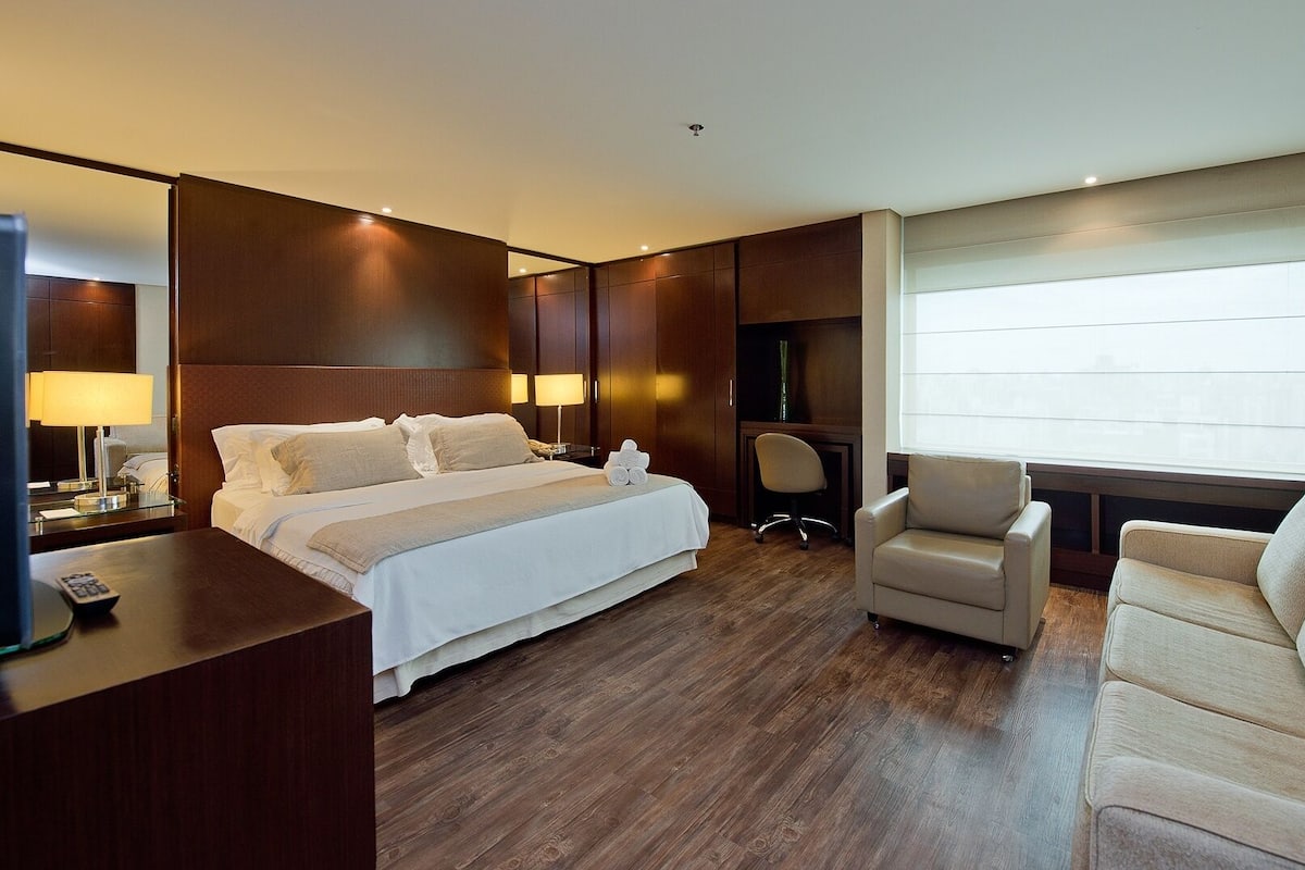 A hotel room with a large bed, a desk with a lamp, a chair, and a sofa.