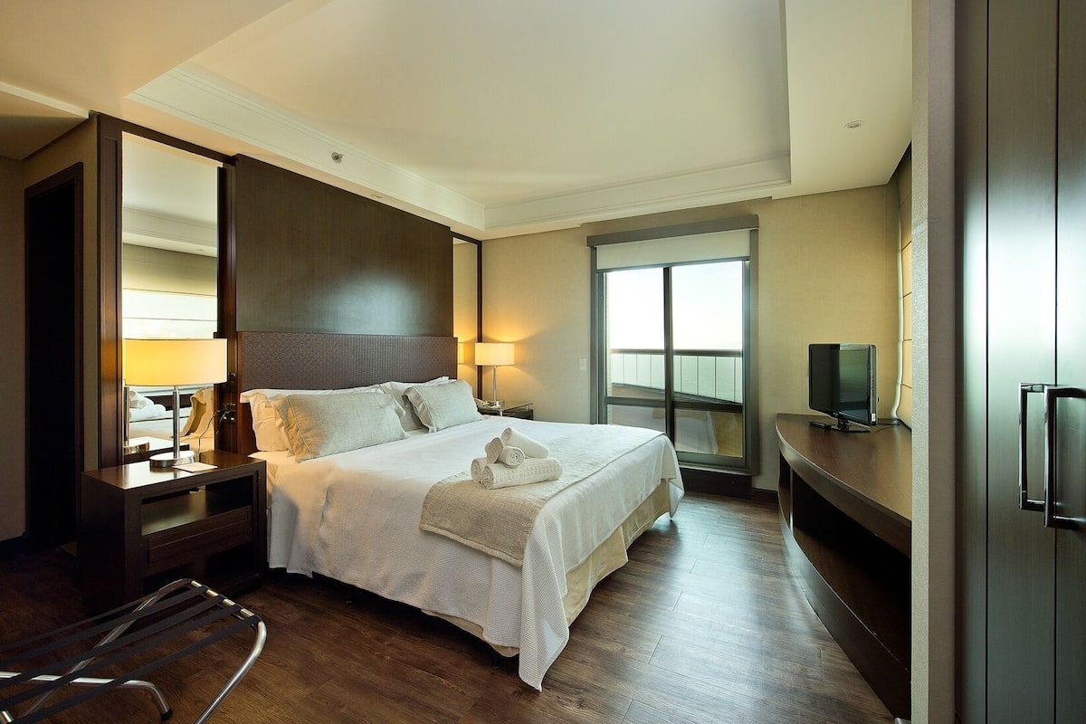 A hotel room with a large bed, a TV, a desk, and a window with a view.