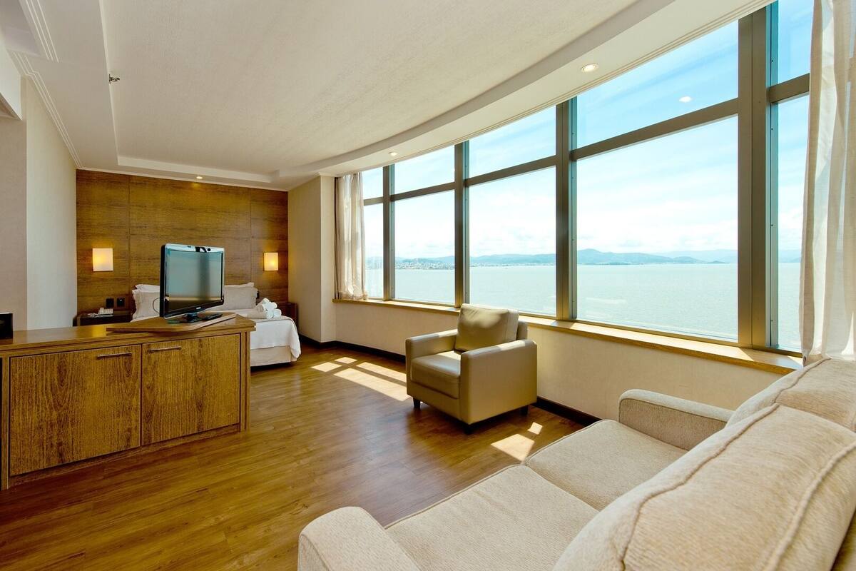 A modern hotel room with a wooden desk, a flat-screen TV, a comfortable sofa, and a large window offering a view of the water.