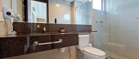 Un baño moderno con cabina de ducha de vidrio, un inodoro blanco y encimera de mármol con secador de pelo y otros artículos de aseo.