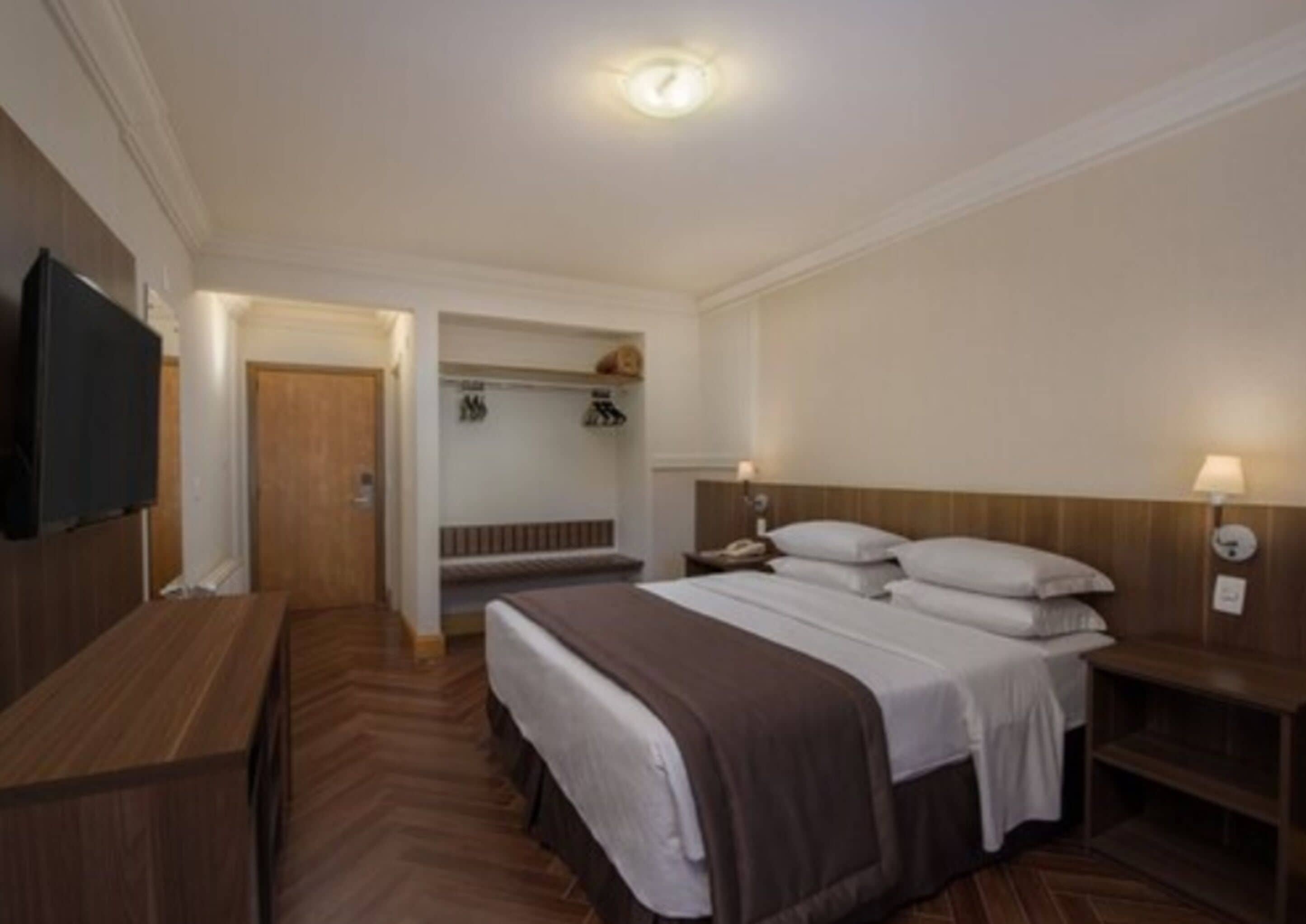 Quarto de hotel com cama, mesas de cabeceira, televisão e piso de madeira.