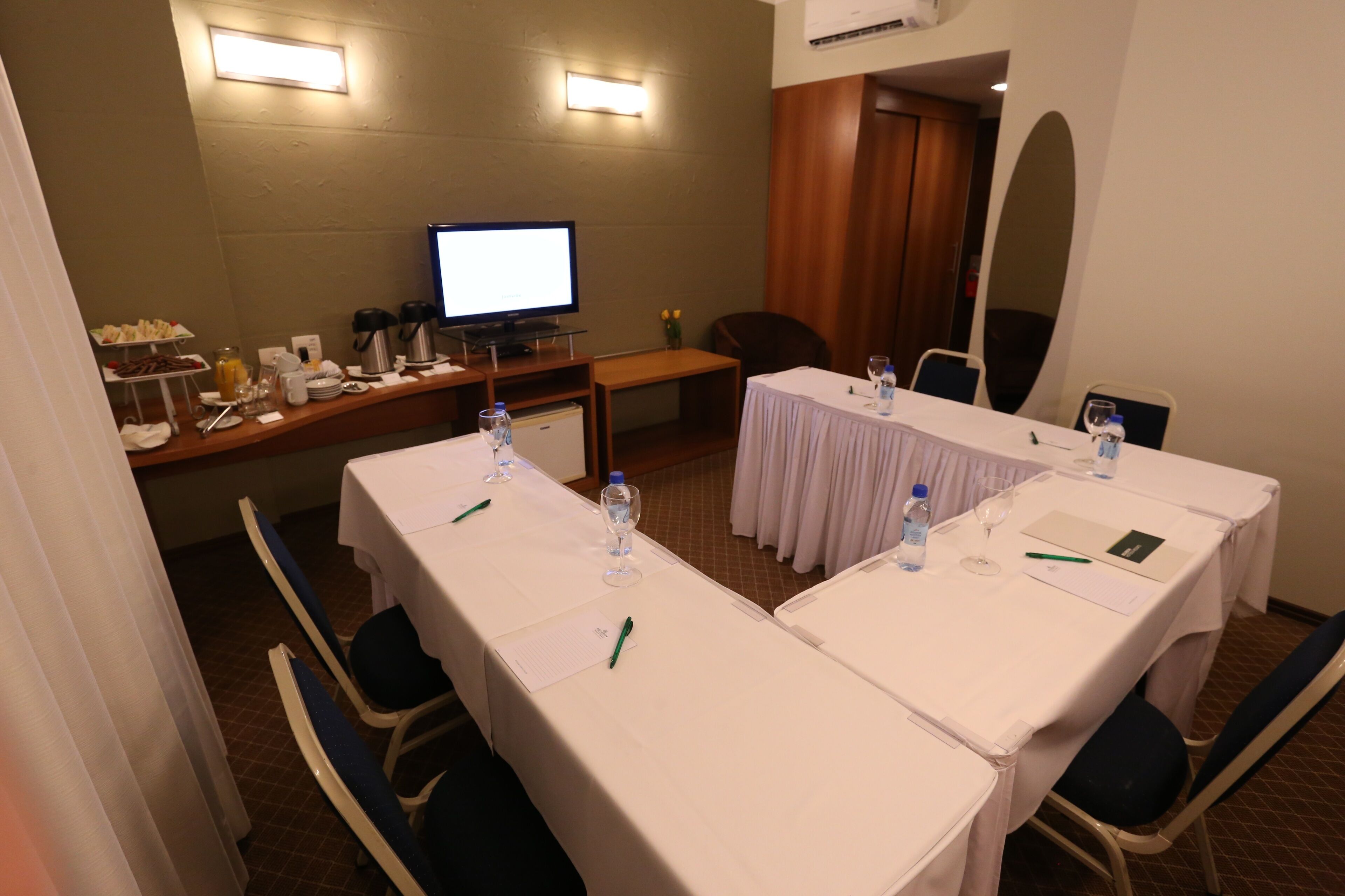 Un salón de conferencias con dos mesas, sillas, un televisor y una zona de buffet con aperitivos.