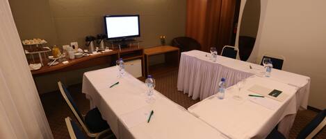 Un salón de conferencias con dos mesas, sillas, un televisor y una zona de buffet con aperitivos.