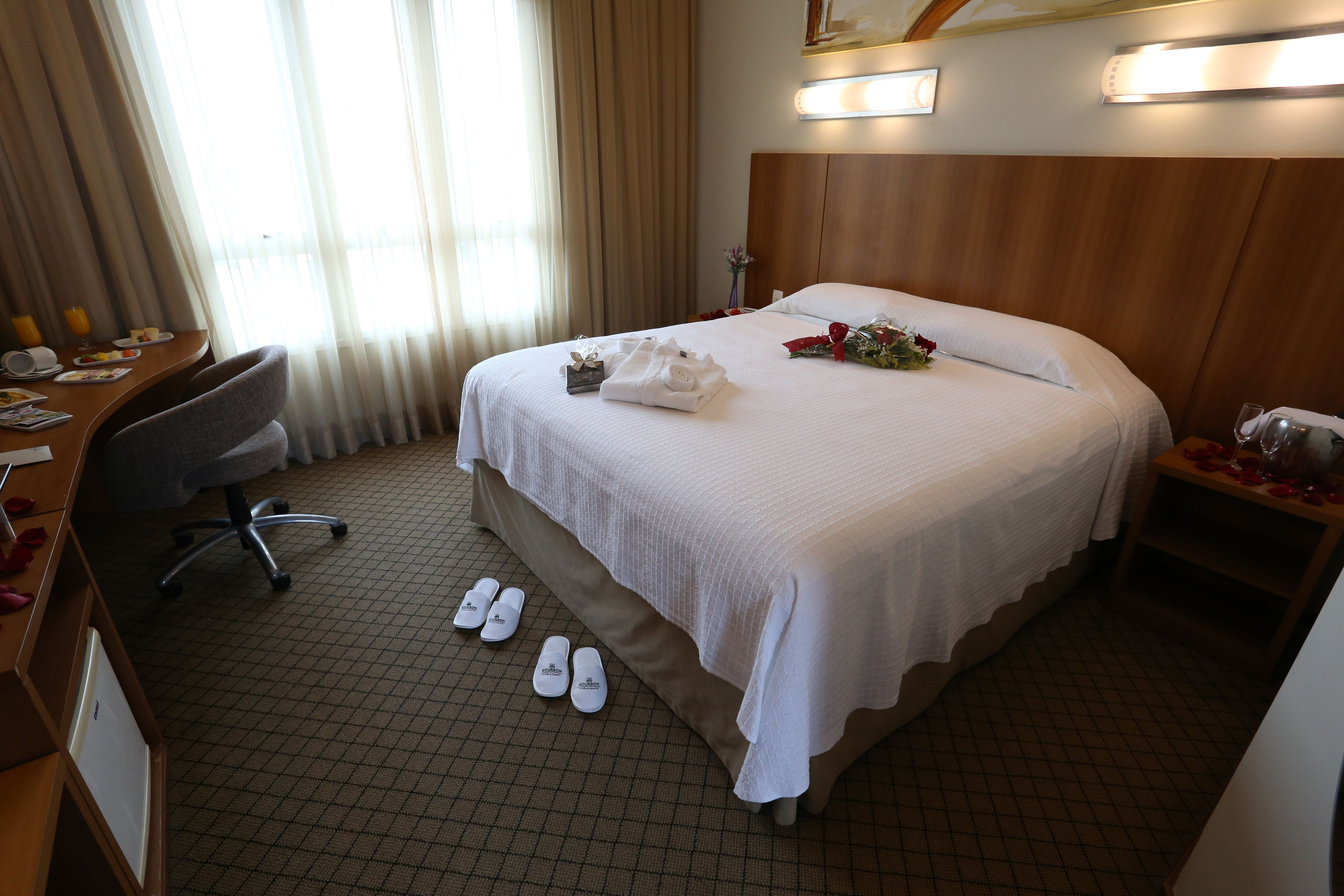 Quarto de hotel com cama arrumada, uma escrivaninha com cadeira, uma mesinha de centro com um vaso, e chinelos no chão.