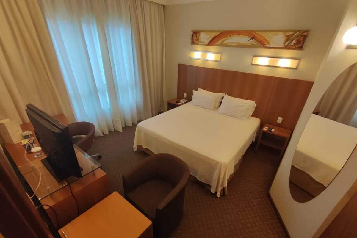 A hotel room with a large bed, a desk with a computer, a chair, a desk lamp, and a mirror.