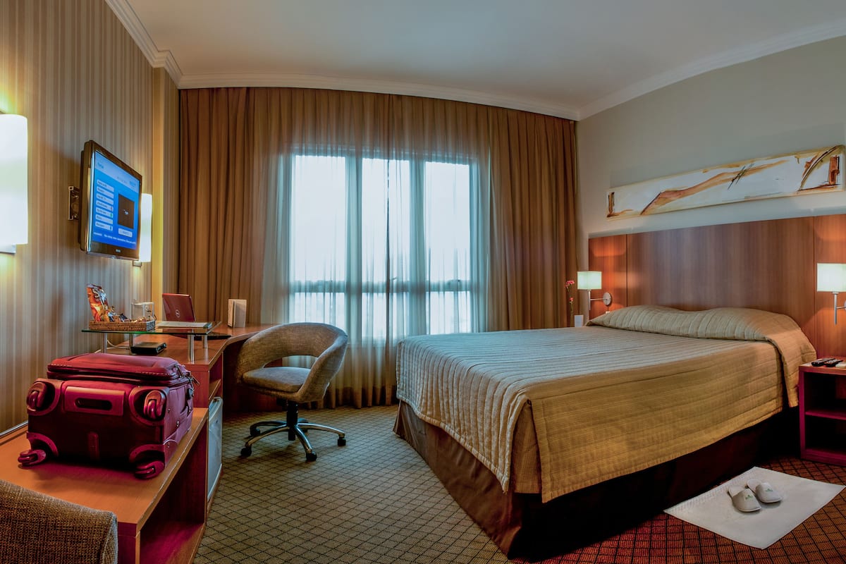 A hotel room with a bed, a desk with a computer, a chair, a TV, and a window with curtains.