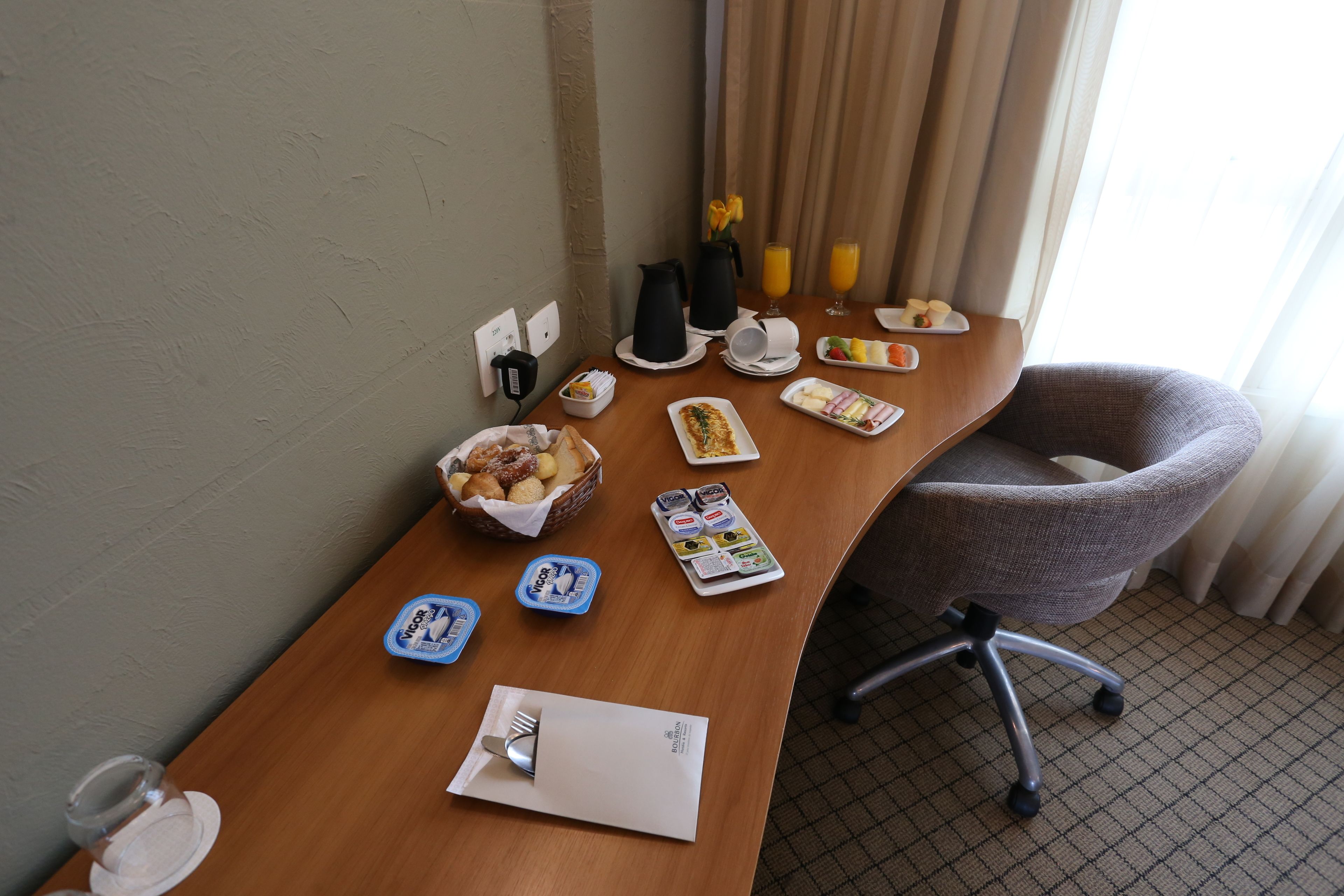 Quarto de hotel com escrivaninha de madeira, itens de café da manhã, uma cadeira e uma janela com cortinas.