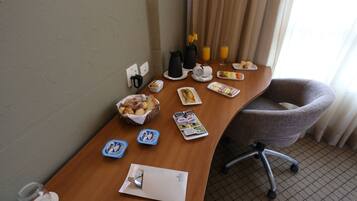 Quarto de hotel com escrivaninha de madeira, itens de café da manhã, uma cadeira e uma janela com cortinas.