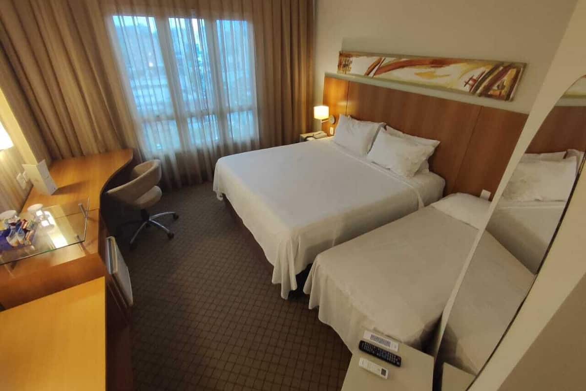 A hotel room with two beds, a desk, a chair, a lamp, and a painting on the wall.