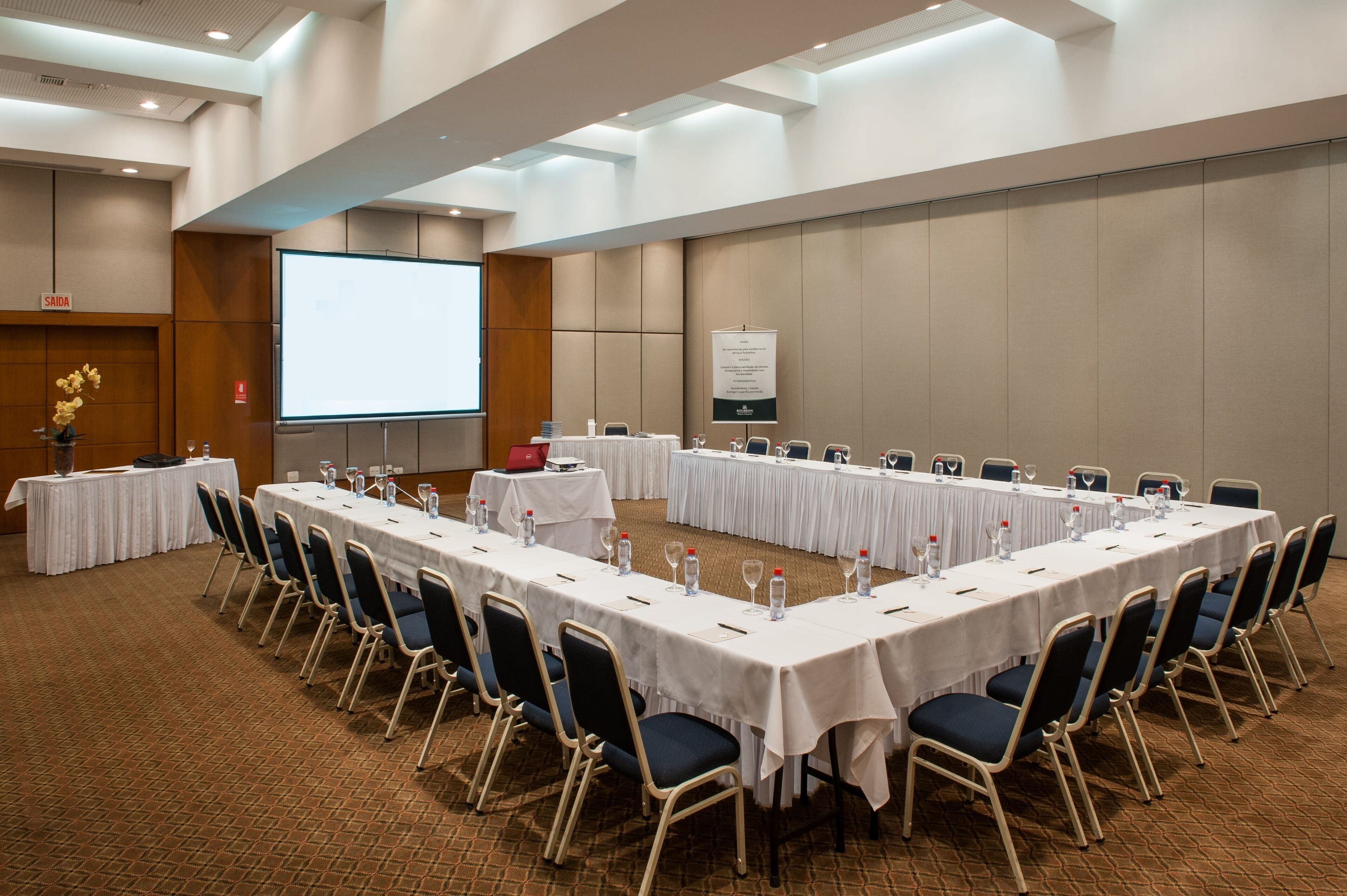 Un salón de conferencias con mesas largas, sillas y una pantalla para proyector.