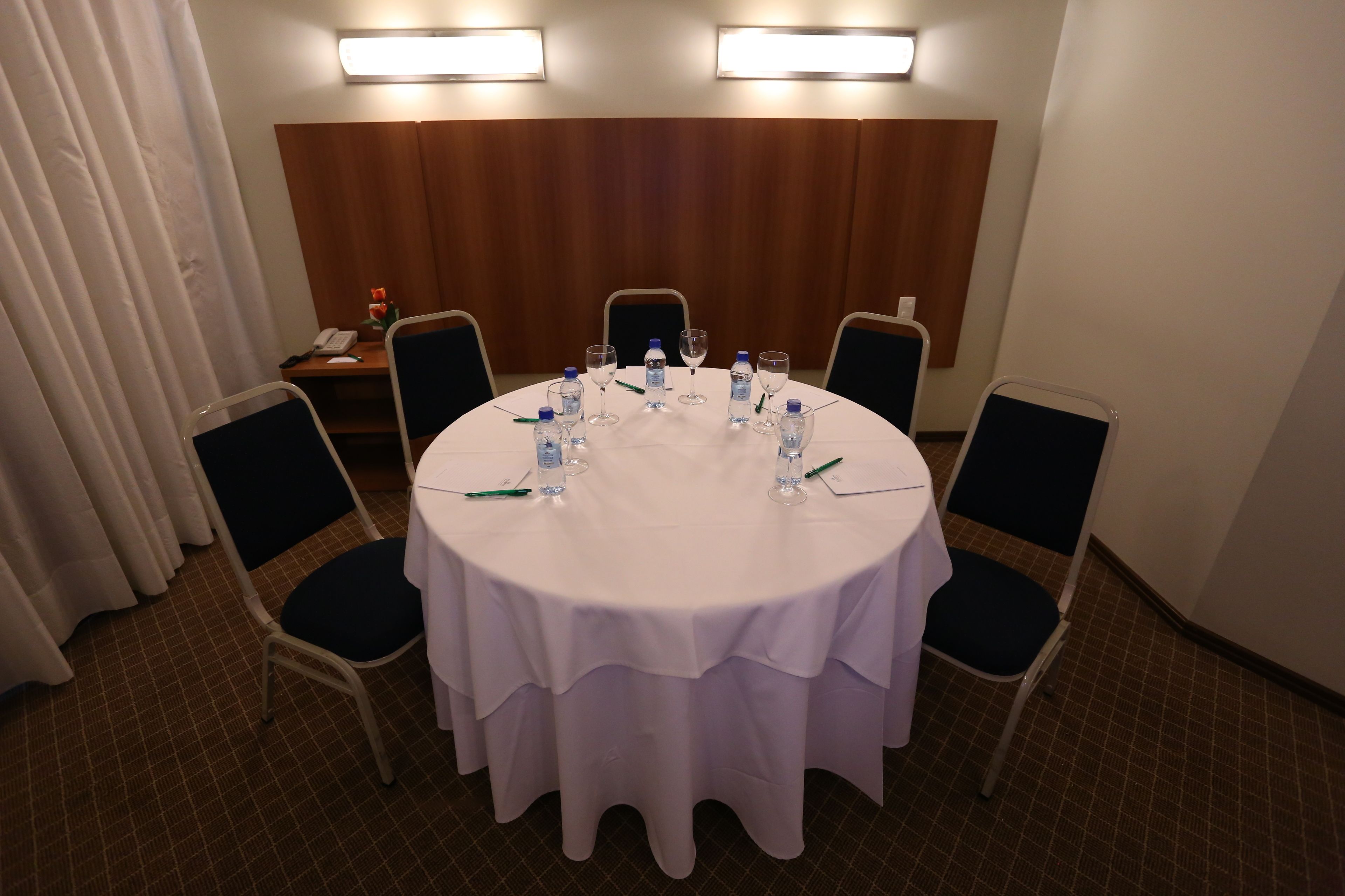 Un salón de conferencias con una mesa redonda, sillas, botellas de agua y un teléfono.
