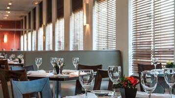 A restaurant interior with tables set for dining, large windows with blinds, and a modern ceiling design.