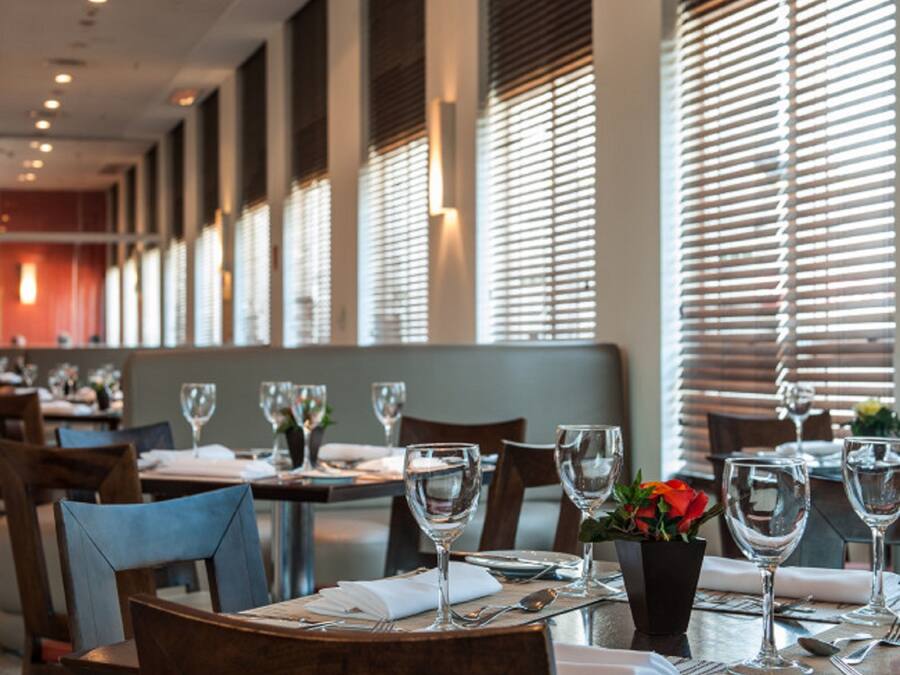 A restaurant interior with tables set for dining, large windows with blinds, and a modern ceiling design.