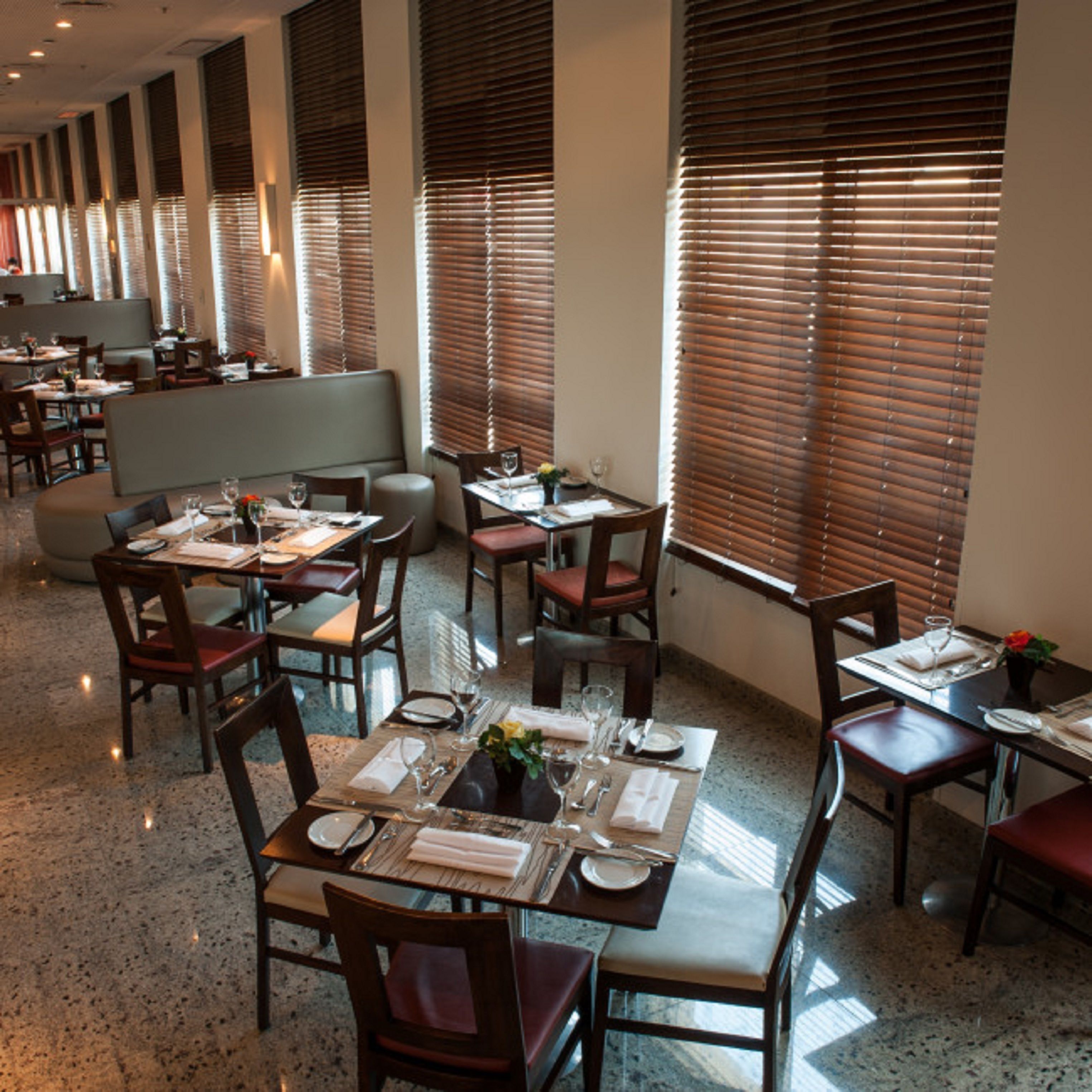 Een restaurant met meerdere tafels en zitjes, gerangschikt in een rij langs een gang met grote ramen en zonwering.