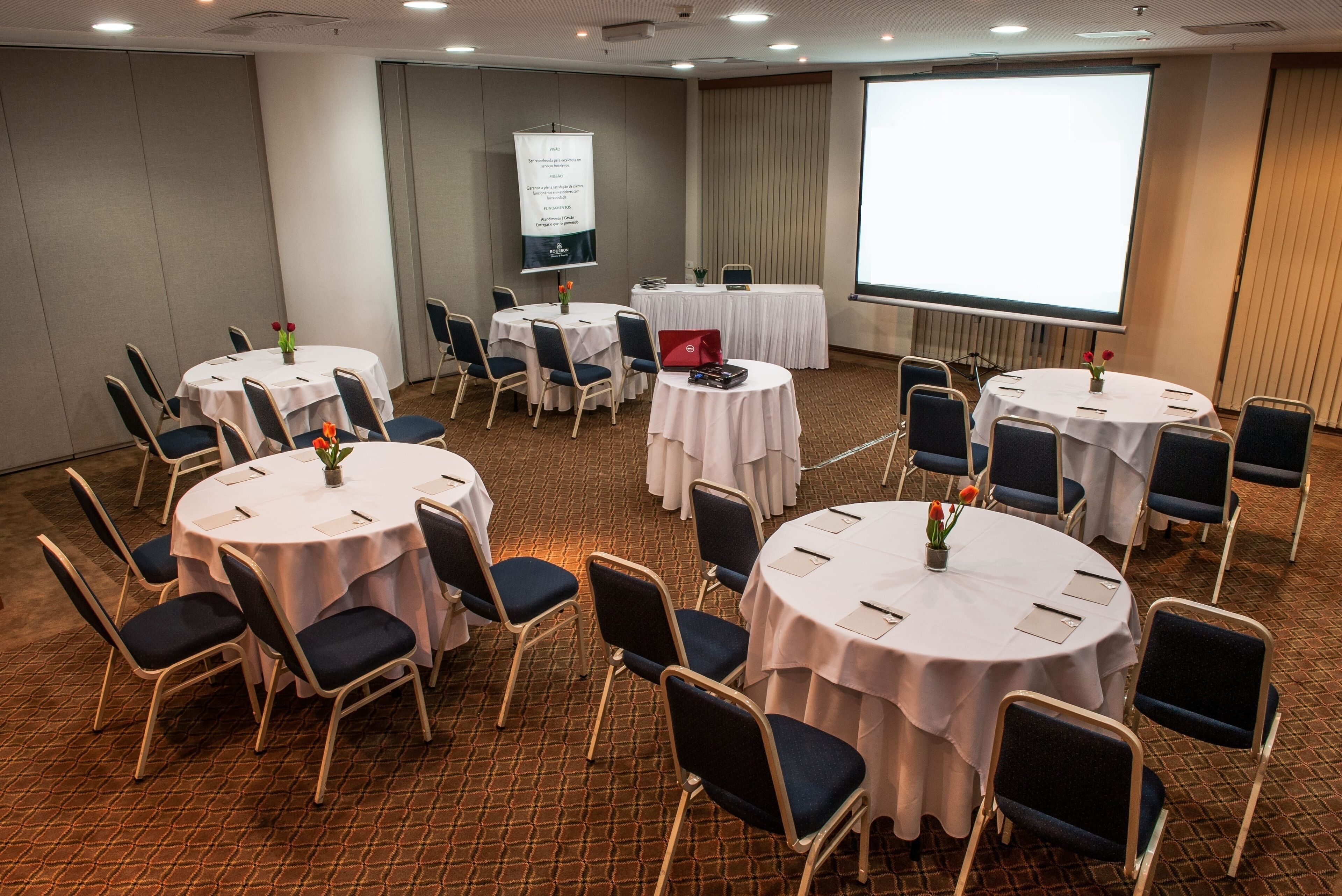 A conference room with multiple round tables, each with a white tablecloth and blue chairs. There is a projector screen and a presentation board on the wall.