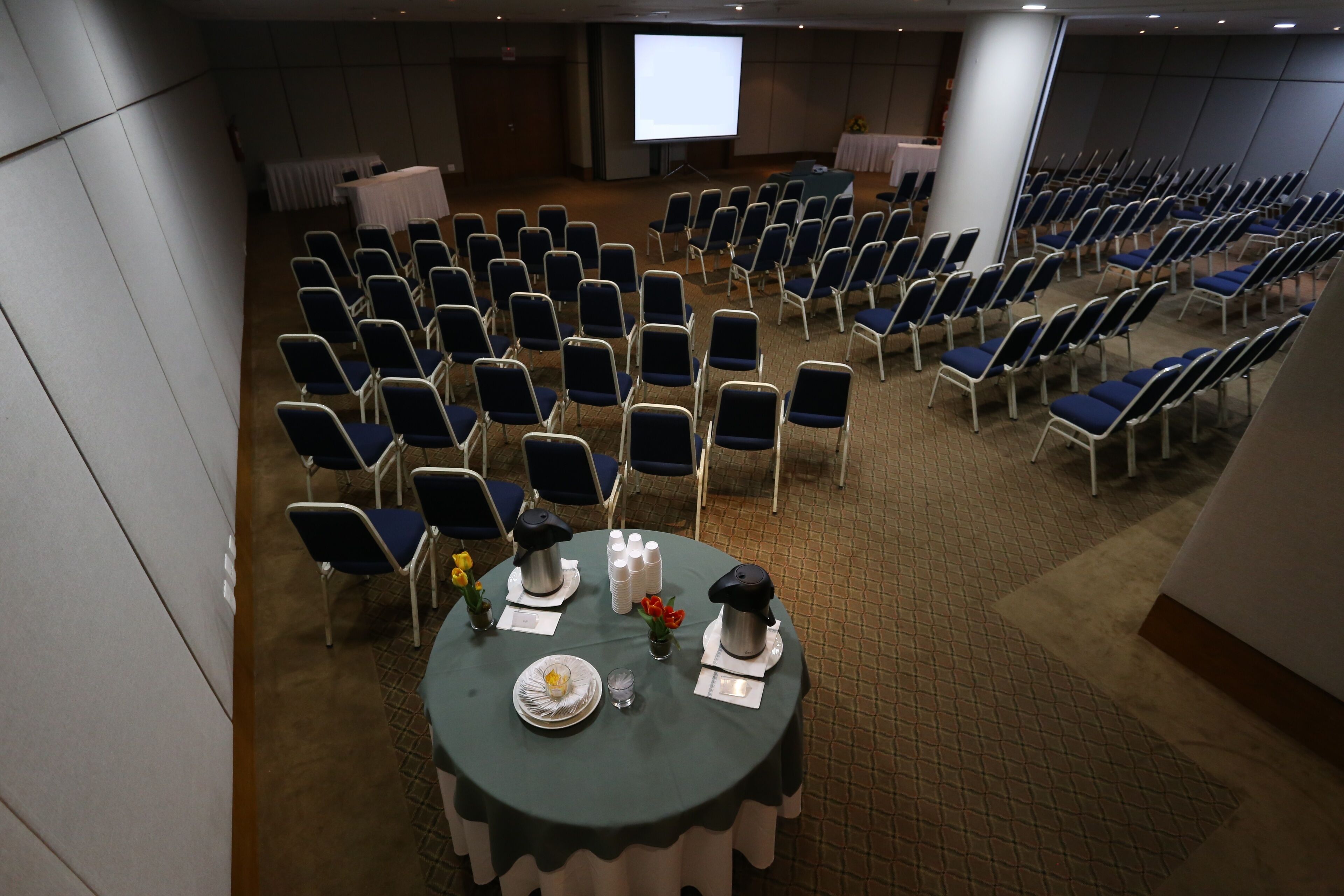 A conference room with rows of chairs, a projector screen, and a table with a floral arrangement and a coffee pot.