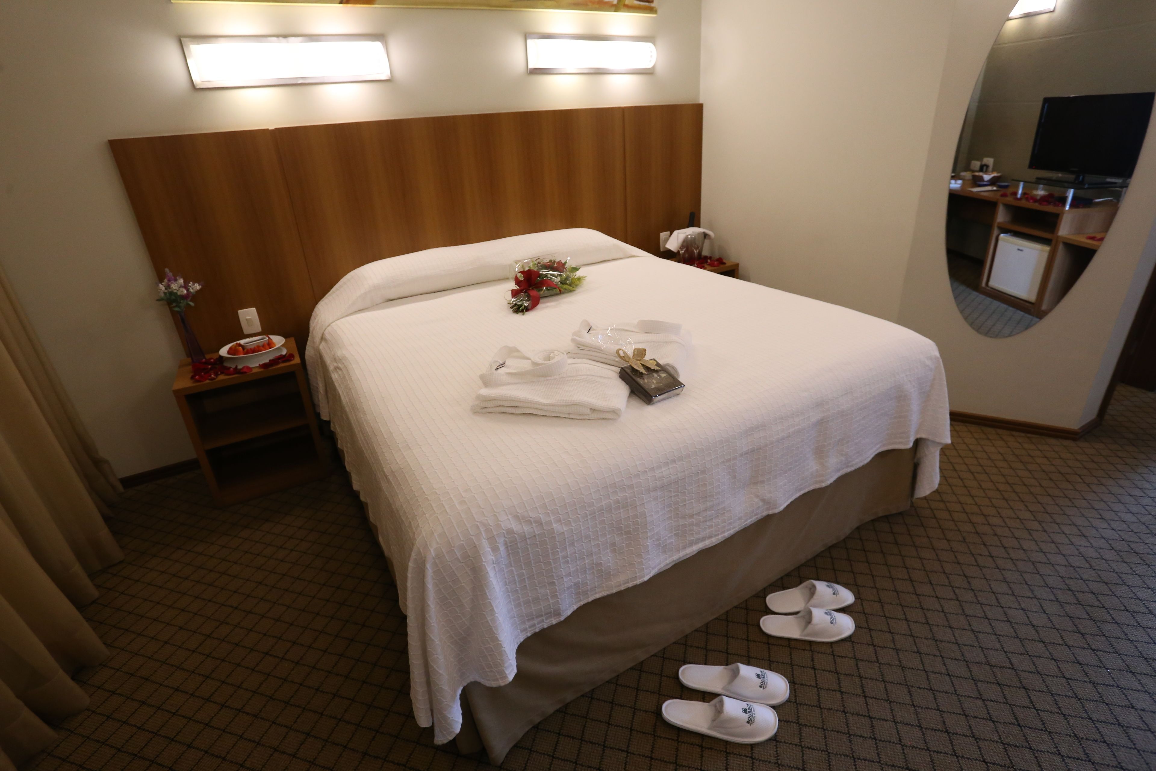 Une chambre d’hôtel avec un grand lit, une tête de lit en bois, une table de chevet ornée d’un vase de fleurs, un miroir et des chaussons posés sur le sol.