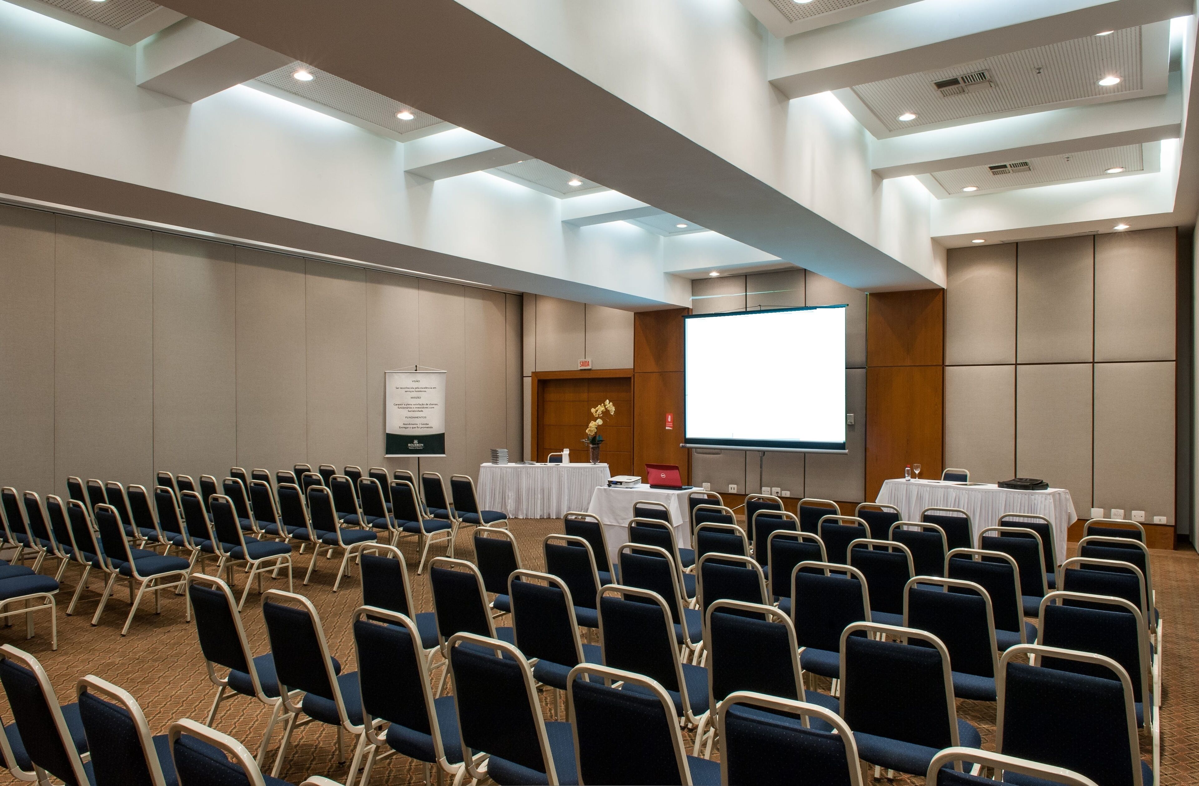 A conference room with rows of chairs, a projection screen, a whiteboard, and tables with nameplates.