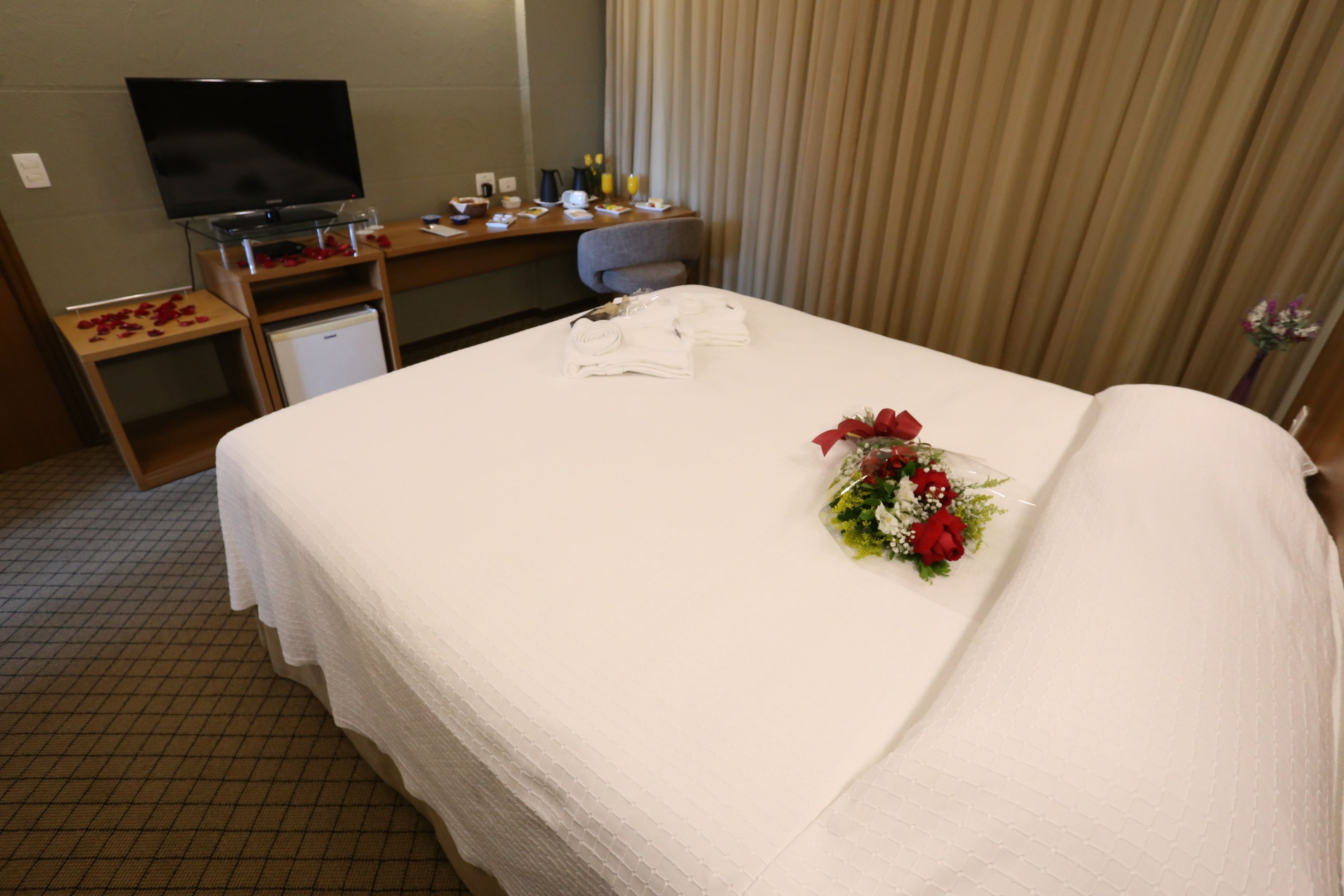 Quarto de hotel com cama, escrivaninha com computador, televisão e pequeno frigorífico.