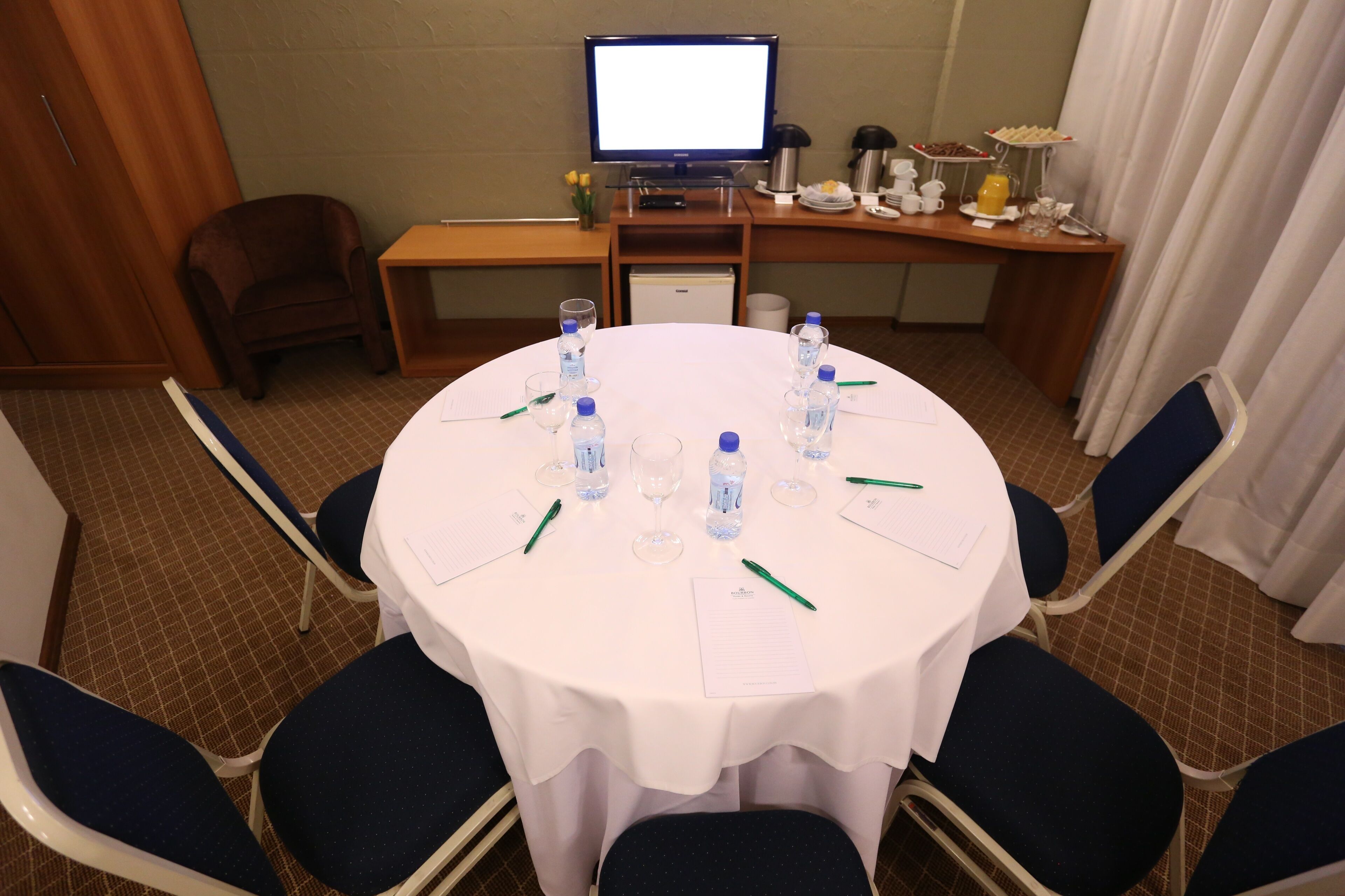 Uma sala de conferências com uma mesa redonda, cadeiras, uma televisão e um buffet com comida e bebidas.