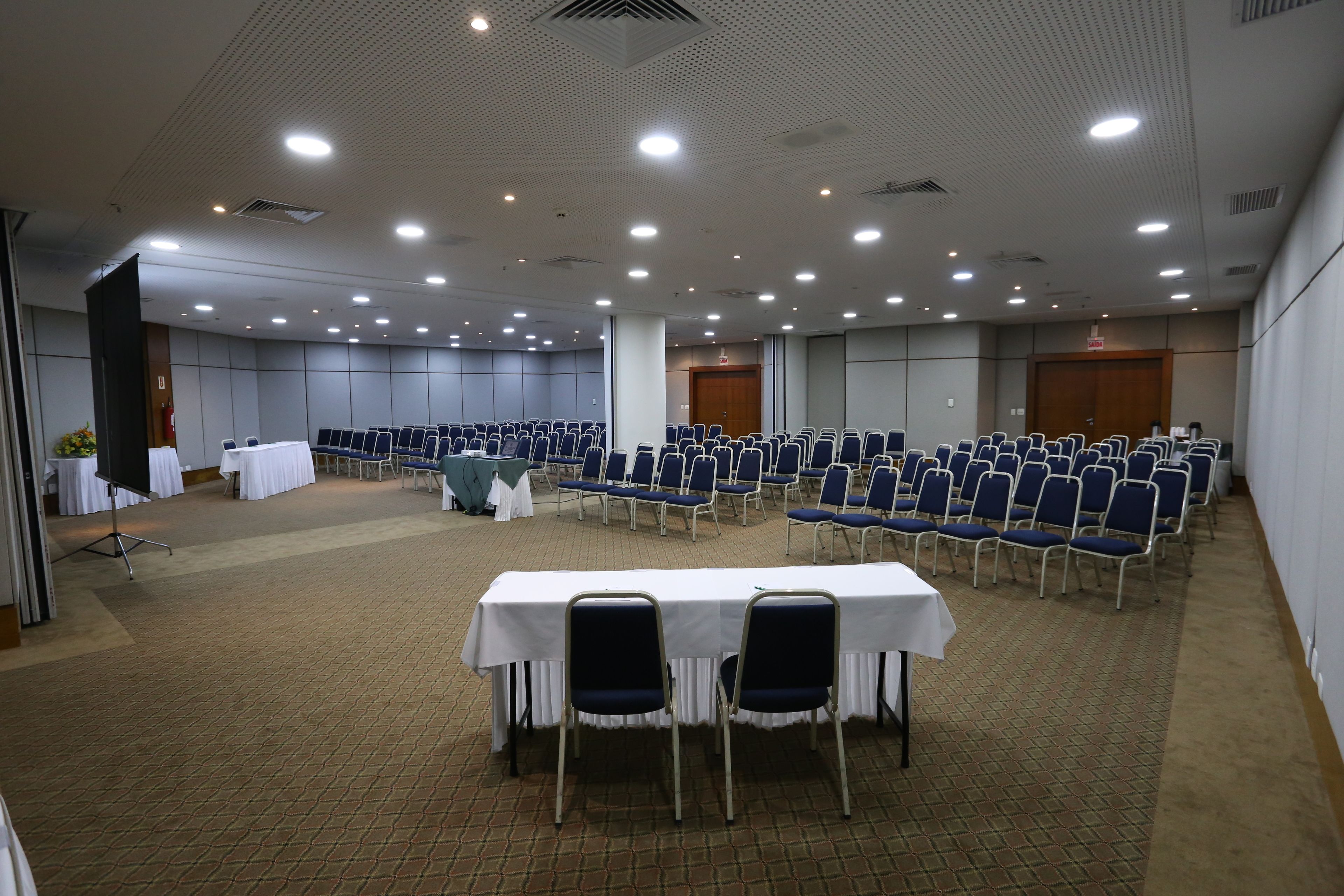 A conference room with rows of blue chairs and white tables, equipped with ceiling lights and a carpeted floor.