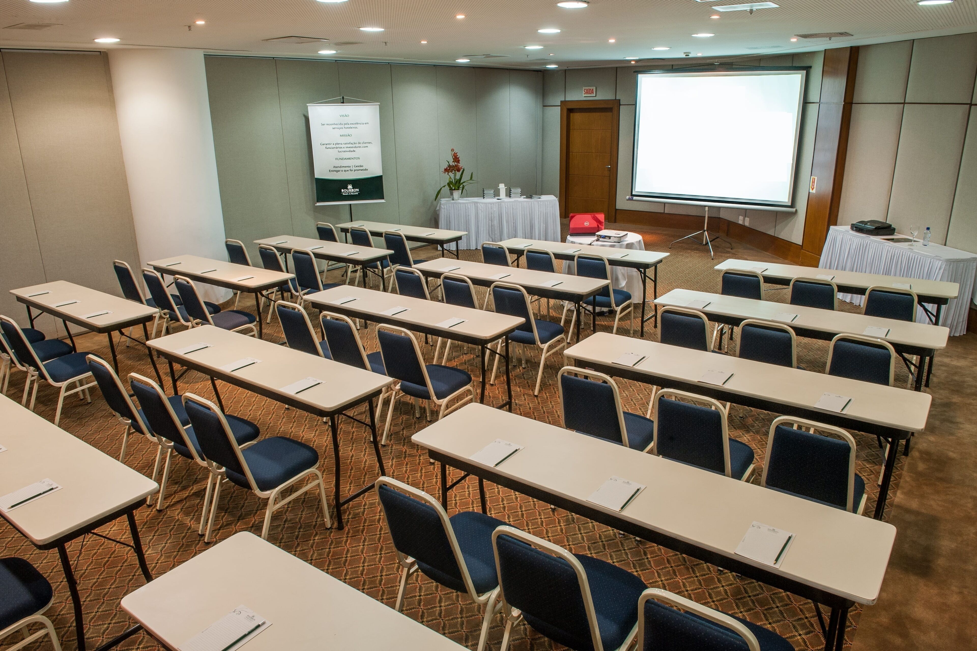 A conference room with rows of tables and chairs, a projector screen, and a whiteboard.