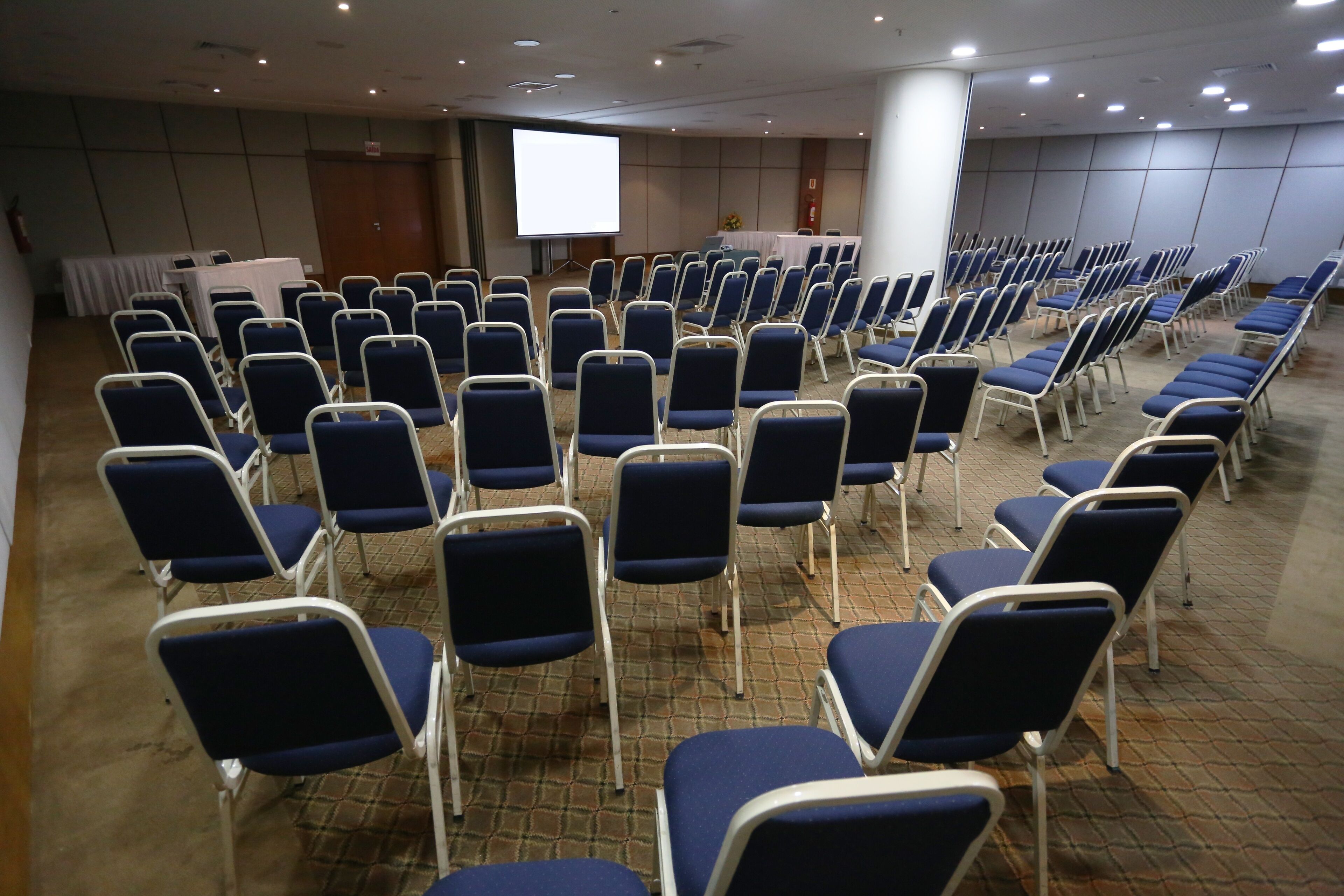 Un salón de conferencias con filas de sillas azules, una pantalla de proyección blanca y una mesa con mantel blanco.