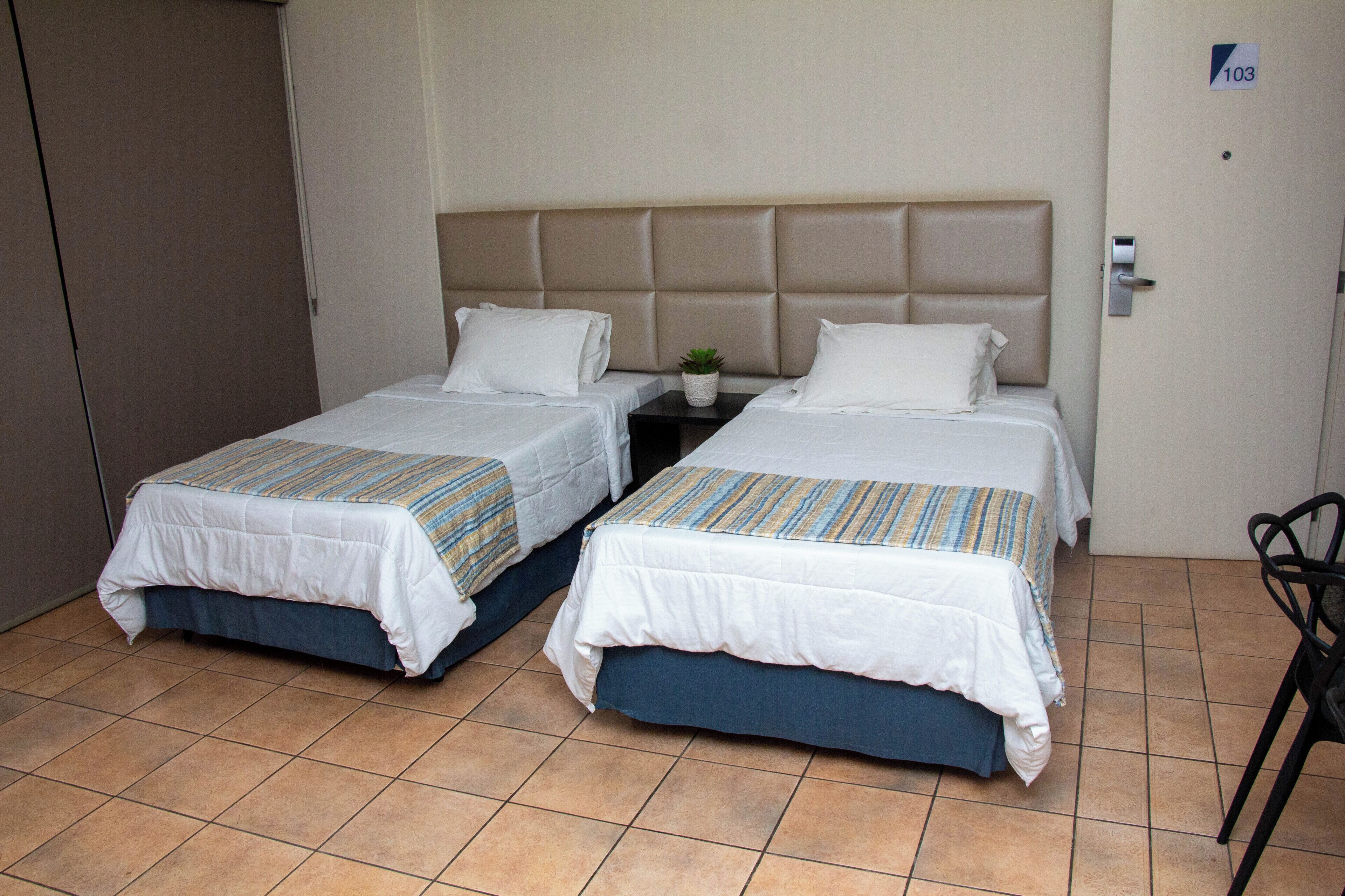 Habitación de hotel con dos camas, una mesita con una planta y una puerta con el número 103.