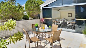 Un área de comedor al aire libre, con paredes de vidrio, sillas de mimbre y una mesa redonda de vidrio, rodeada de plantas en macetas y un patio con baldosas.