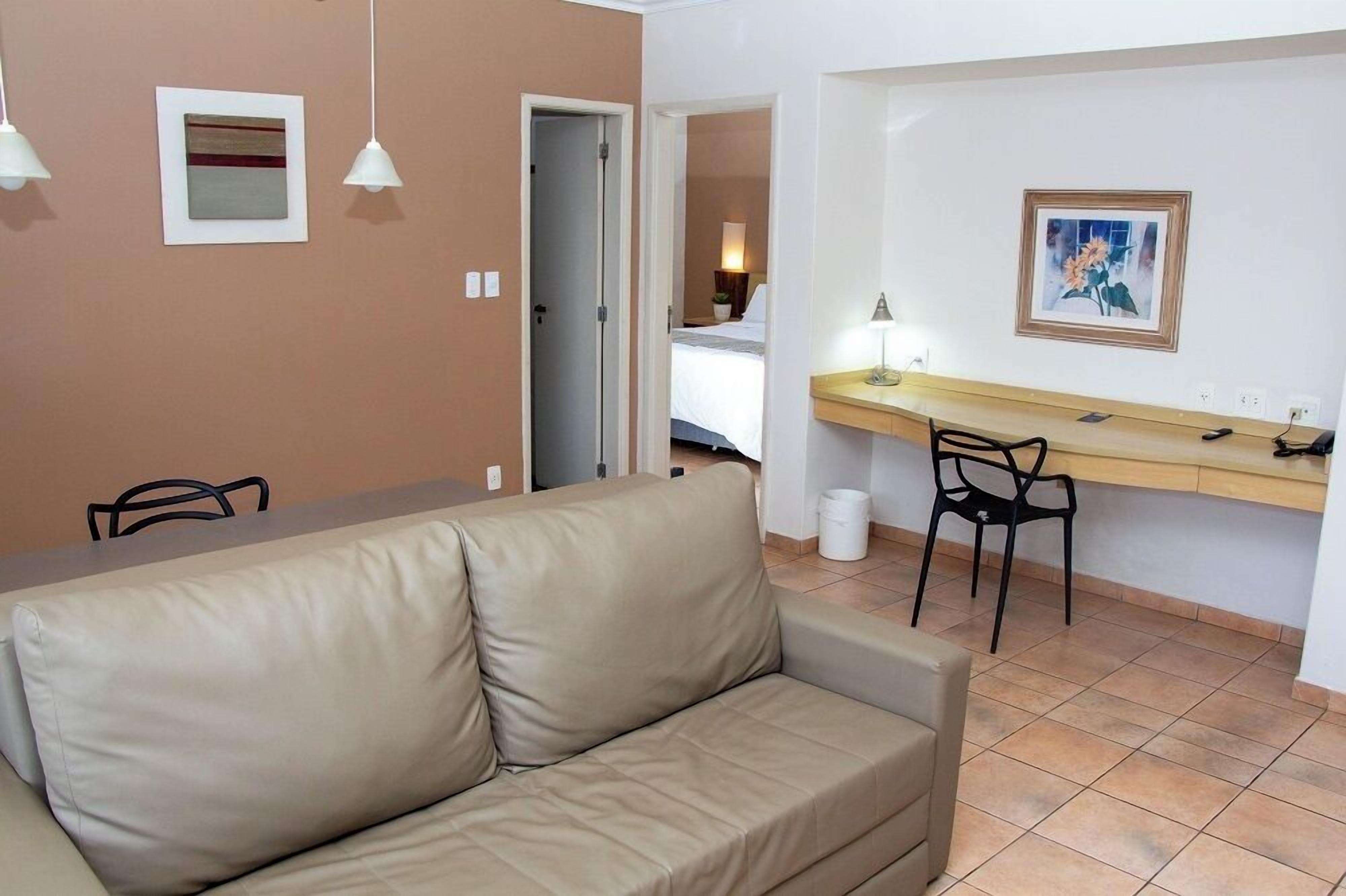 Una habitación con un sofá color beige, un escritorio con una silla, un cuadro en la pared y una puerta que da a otra habitación.