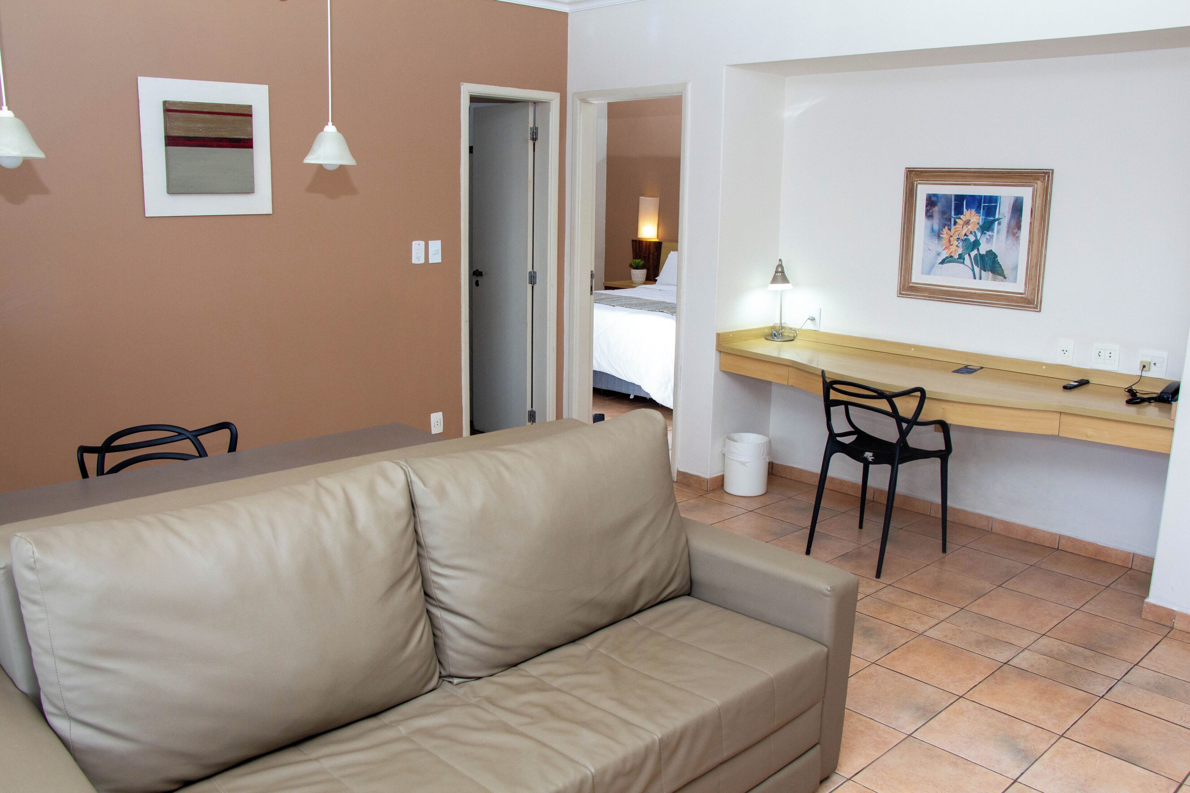 Una habitación con un sofá color beige, un escritorio de madera, una silla y una cama al fondo.