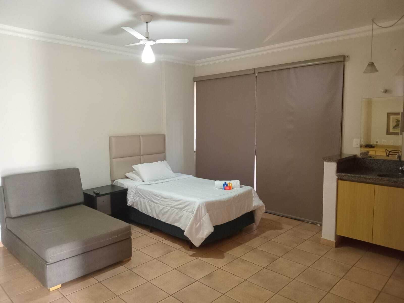 A bedroom with a bed, a small sofa, a nightstand, a ceiling fan, a window with a blind, and a kitchenette area with a sink and countertop.