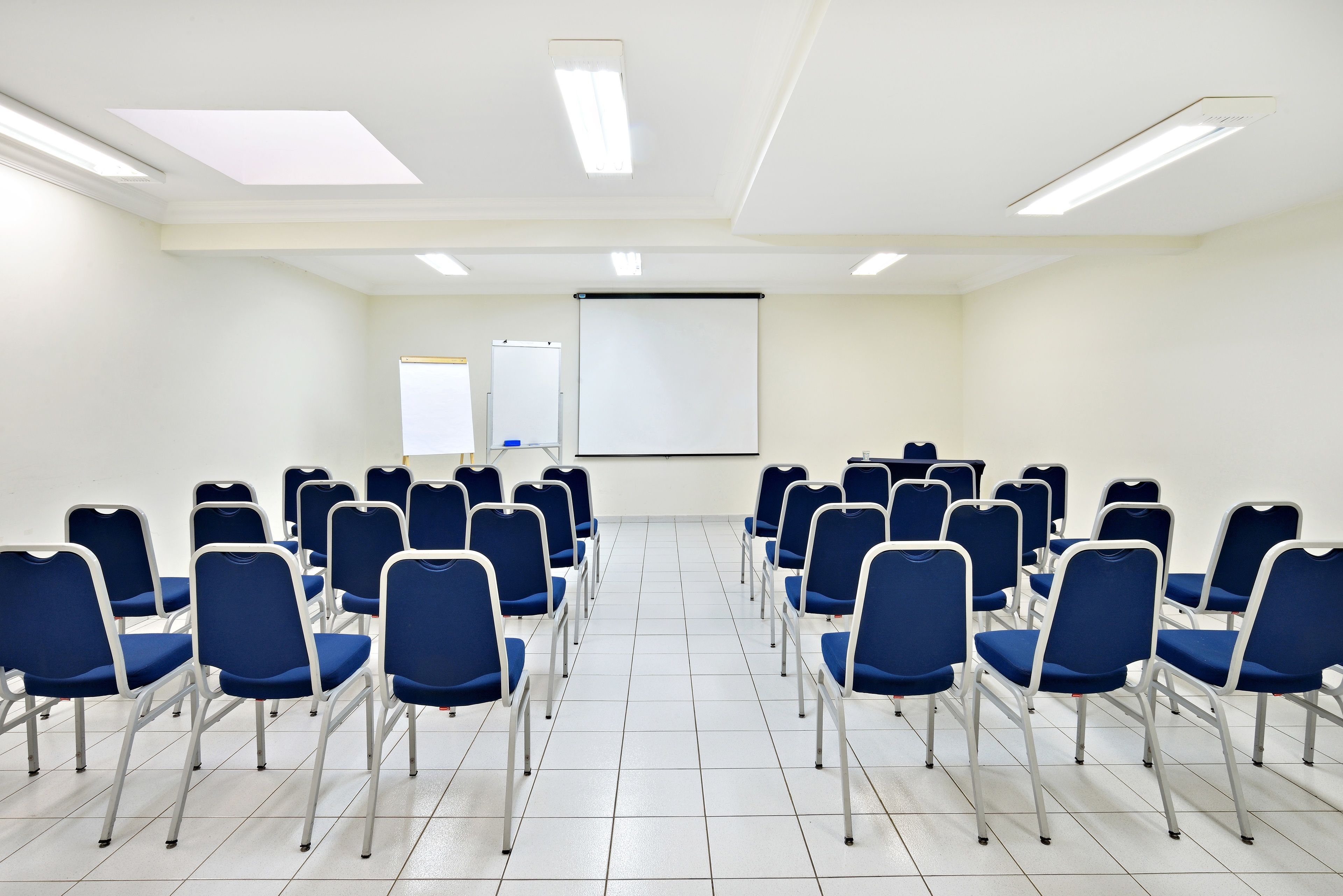 Un salón de conferencias con filas de sillas azules, una pantalla de proyector y una pizarra.