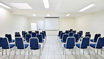 Un salón de conferencias con filas de sillas azules, una pantalla de proyector y una pizarra.