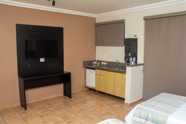 Habitación de hotel con televisor de pantalla plana, kitchenette con lavamanos y armarios, y cama con colcha a rayas.