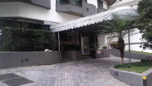 Un edificio moderno con acceso cubierto, paseo pavimentado y zonas ajardinadas con plantas.